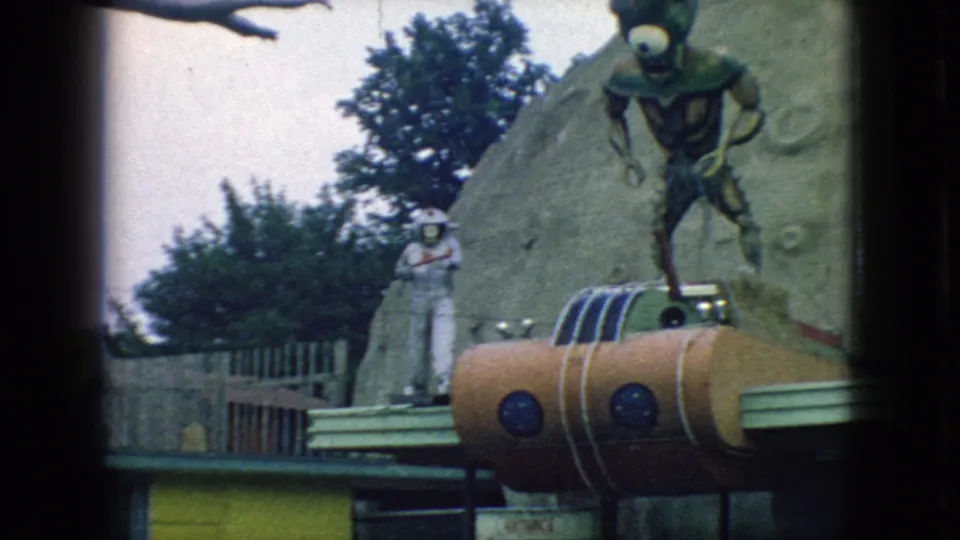 1966 New Jersey 8mm frame showing the astronaut figure more clearly beside the alien and orange rocket structure, with fencing and trees in the background.