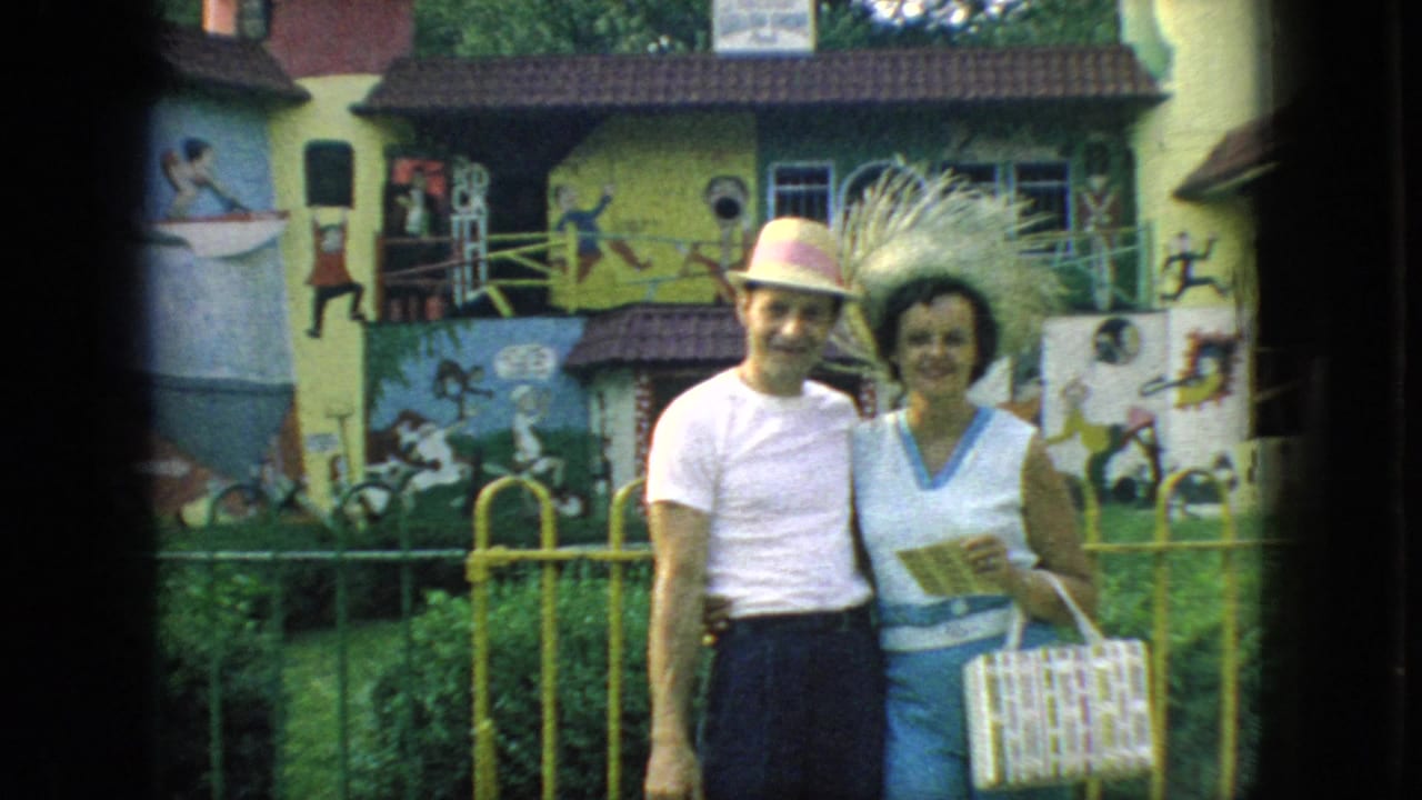 1966 New Jersey home-movie frame of an adult man and woman standing together before a yellow fence and brightly painted mural-covered building.