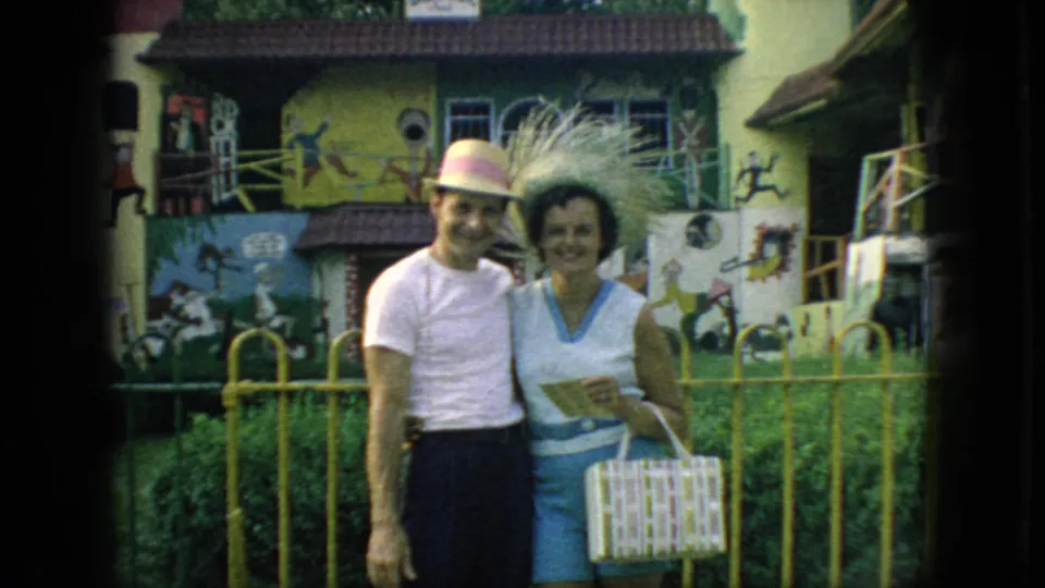 1966 New Jersey 8mm frame showing an adult couple smiling close to the camera, with a yellow fence, painted figures, and a patterned purse visible.