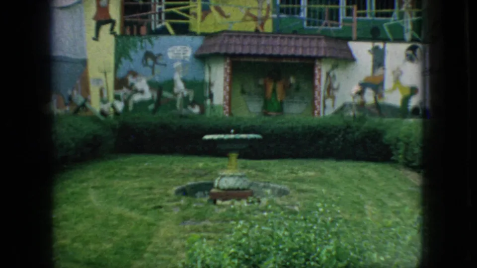1966 New Jersey home-movie frame showing a small fountain and trimmed lawn before a brightly painted building with cartoon-like wall murals.