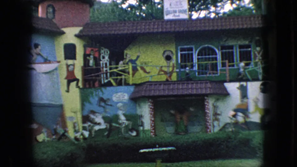 1966 New Jersey 8mm frame showing the mural building more tightly, with painted figures, a balcony-like railing, arched windows, roof tiles, and a low hedge in front.