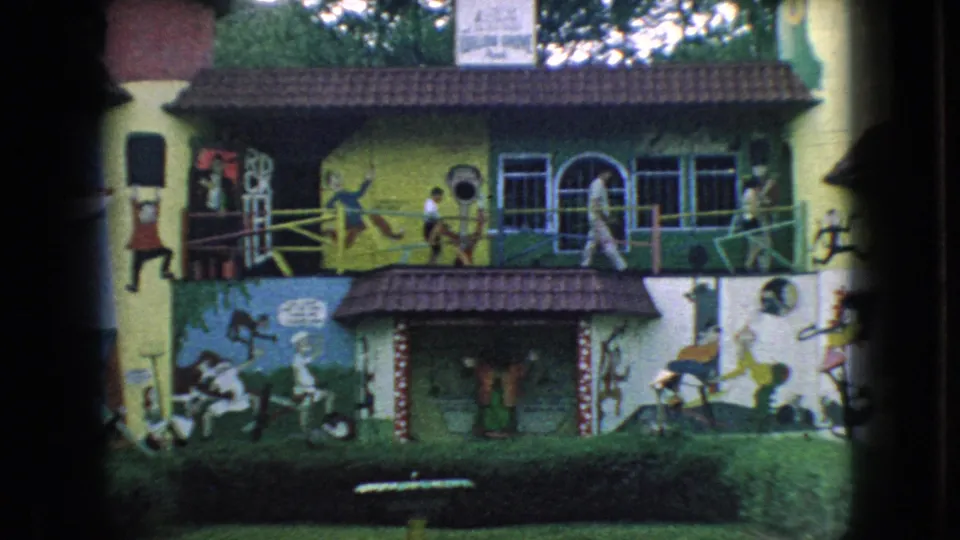 1966 New Jersey 8mm frame showing the colorful mural-covered building, lawn, arched windows, partial lettering, and an unidentified person walking along the front.