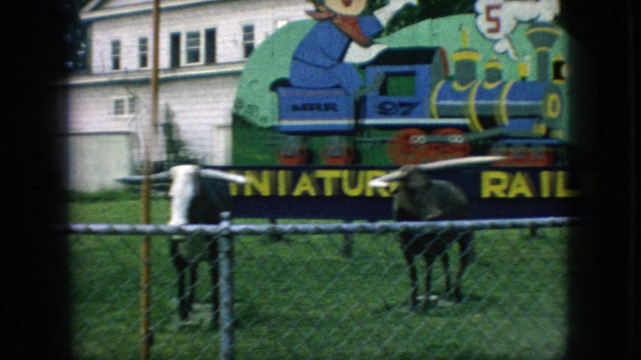 1966 New Jersey home-movie frame with a colorful miniature railway sign, cartoon train artwork, and longhorn statues on grass behind a chain-link fence.