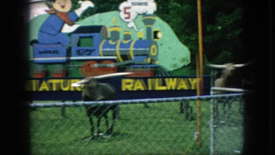 1966 New Jersey 8mm still showing the painted miniature railway sign and train illustration behind a longhorn statue and chain-link fence.