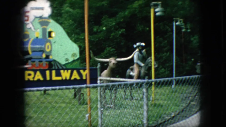 1966 New Jersey frame showing longhorn statues near a chain-link fence, with part of the miniature railway sign and trees in the background.