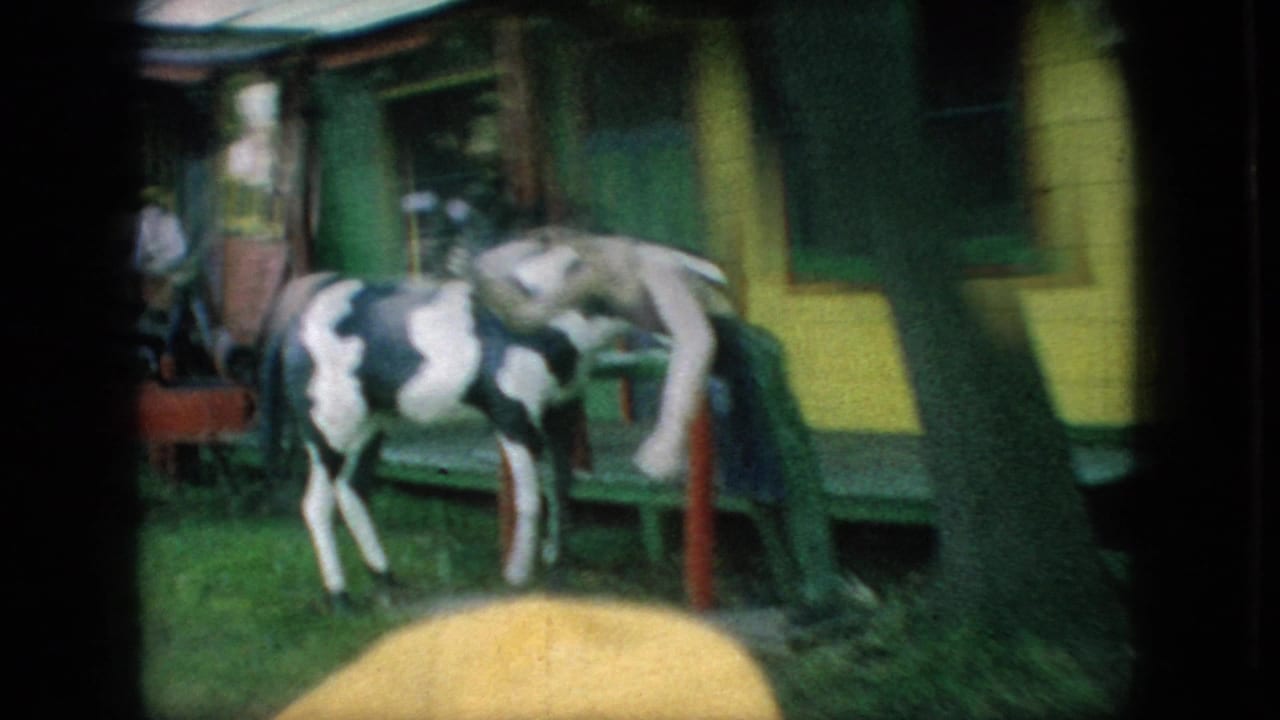 1966 New Jersey 8mm frame showing a black-and-white cow figure near colorful wooden facades and a cowboy-hatted figure at an outdoor amusement attraction.
