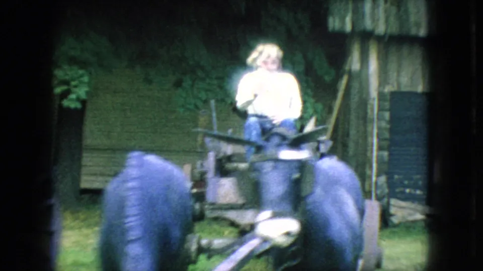 A rider in a light shirt and hat sits on the mule-drawn cart as it passes a rustic exterior wall and grassy yard in 1966 New Jersey footage.