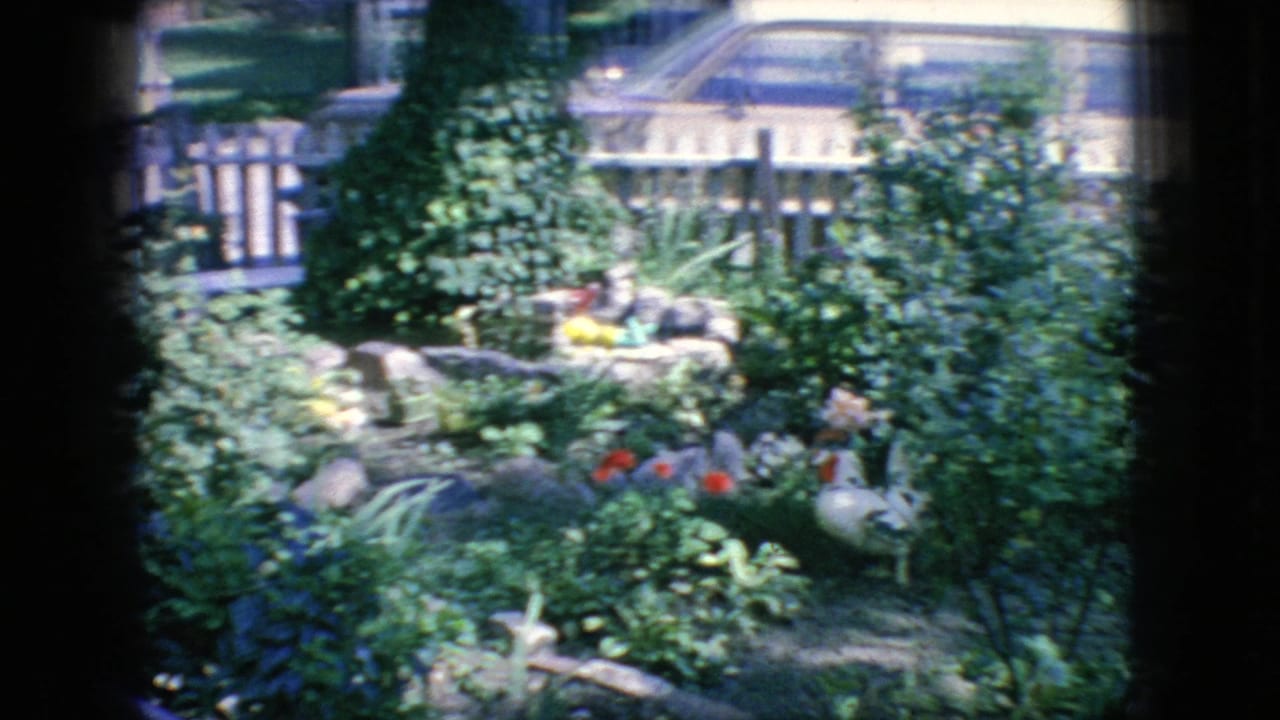 Flower garden with shrubs, rocks, small fountain or ornament, picket fence, and parked cars behind it.