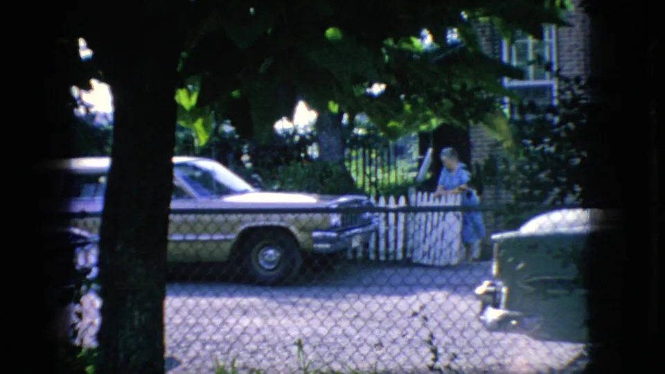 1966 New Jersey home-movie frame showing a woman in a blue dress at a white picket gate, with a classic station wagon, chain-link fence, trees, and a brick house nearby.