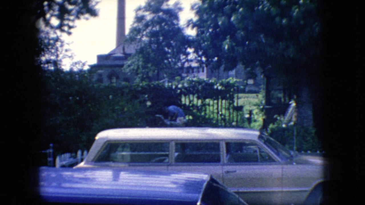 1966 New Jersey home-movie frame with a light-colored station wagon in the foreground, leafy fencing behind it, and a spired building partly visible through trees.