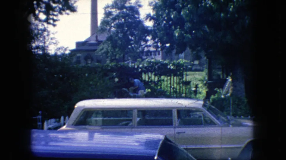 1966 New Jersey home-movie frame showing a station wagon behind a blue car hood, with fencing, dense greenery, and a spired building in the background.