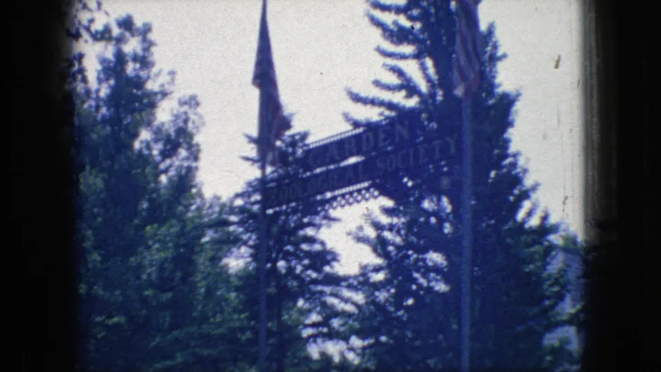 1966 New Jersey home-movie frame showing a partially readable zoo entrance sign among trees, with a tall pointed spire and bright sky in the background.
