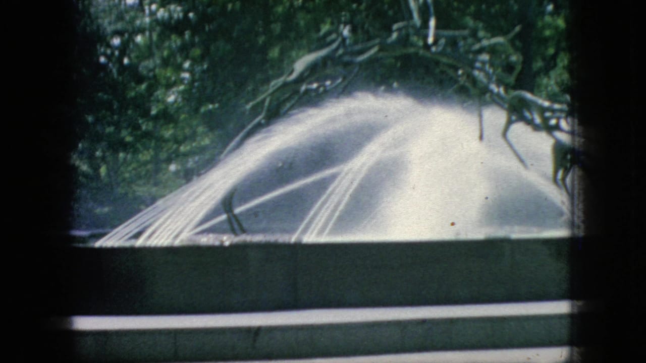 1966 New Jersey home-movie frame showing a water-spraying sculptural fountain with leafy trees behind it, partially obscured by dark foreground framing.