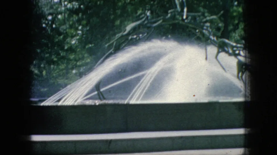 1966 New Jersey home-movie frame showing a water-spraying sculptural fountain with leafy trees behind it, partially obscured by dark foreground framing.