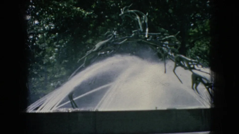 1966 New Jersey home-movie frame of the same outdoor fountain spraying water, with tree cover behind and dark window-like framing at the edges.