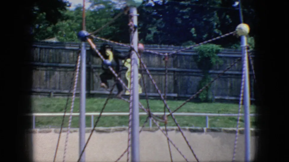 1966 New Jersey 8mm frame showing a monkey on the rope jungle gym with a partly visible person in yellow behind the structure, plus grass, trees, and a wooden fence.