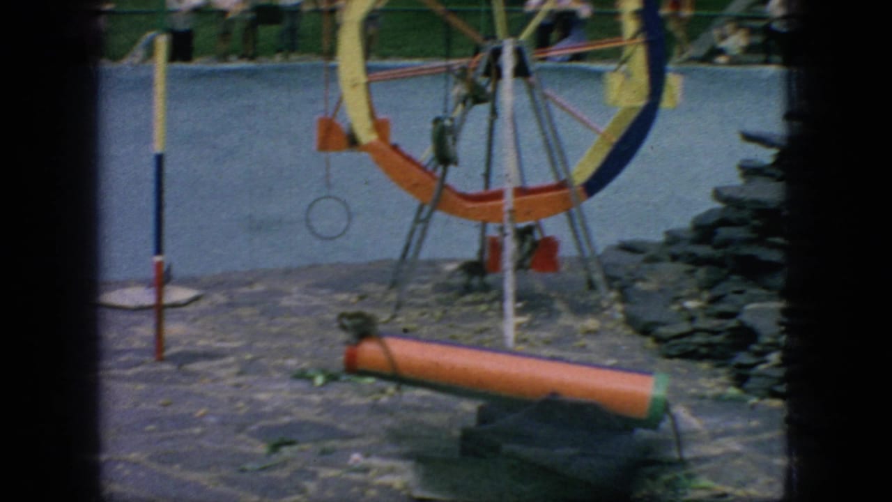 1966 New Jersey 8mm zoo footage showing a monkey near a low orange cylinder, with a painted wheel structure, hanging ring, stone paving, and distant visitors in the background.