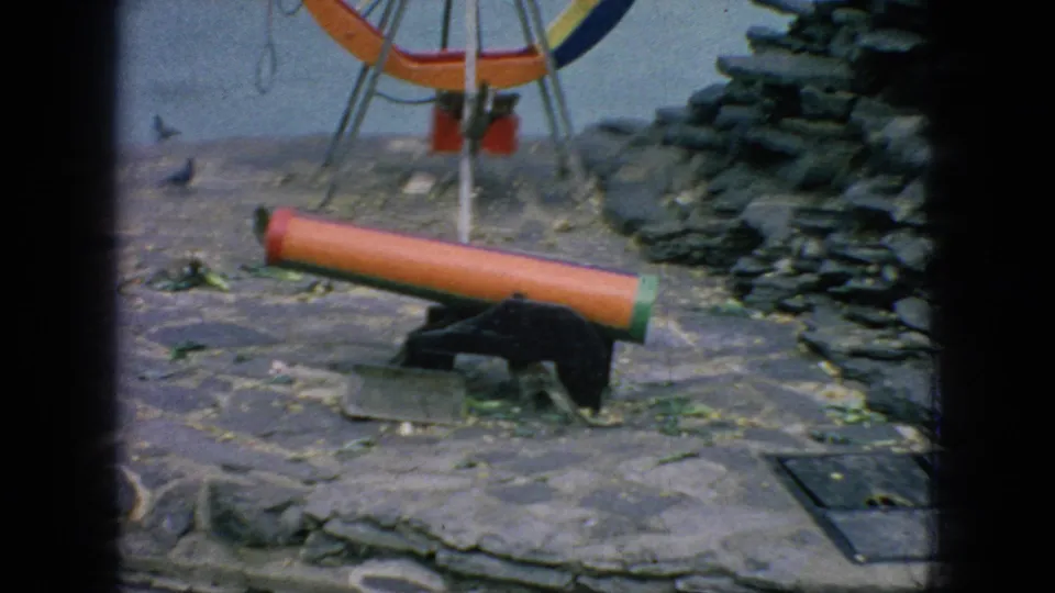 In the final frame, the monkey is near the end of the low orange cylinder on the stone enclosure floor, with the painted wheel apparatus, rock wall, leaves, and a dark access panel visible nearby.