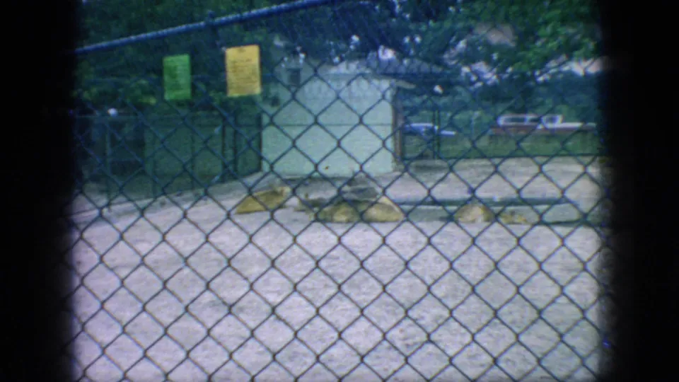 A fenced zoo or animal-park enclosure appears mostly empty in this 1966 New Jersey frame, with chain-link mesh, small signs, trees, and a pale ground surface visible.