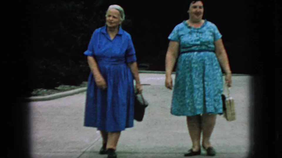 1966 New Jersey home-movie frame of two older women in blue dresses carrying handbags as they walk toward the camera on a paved park walkway.