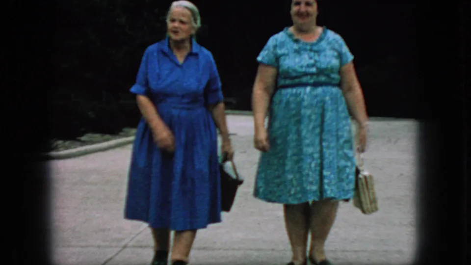 Closer 1966 New Jersey 8mm frame showing two older women in blue dresses and handbags walking together on a tree-lined park path.