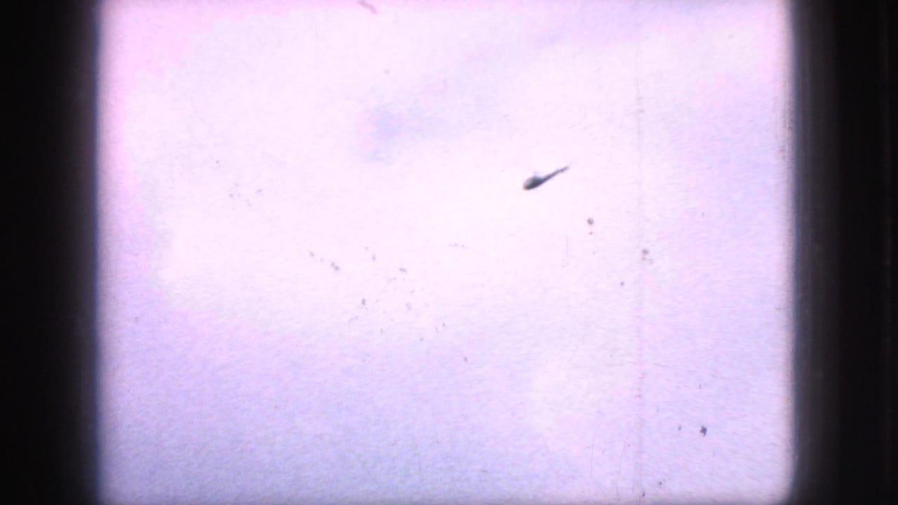 A distant helicopter-like silhouette crosses a pale sky in 1966 New Jersey 8mm footage, framed by dark gate edges and visible film dust.