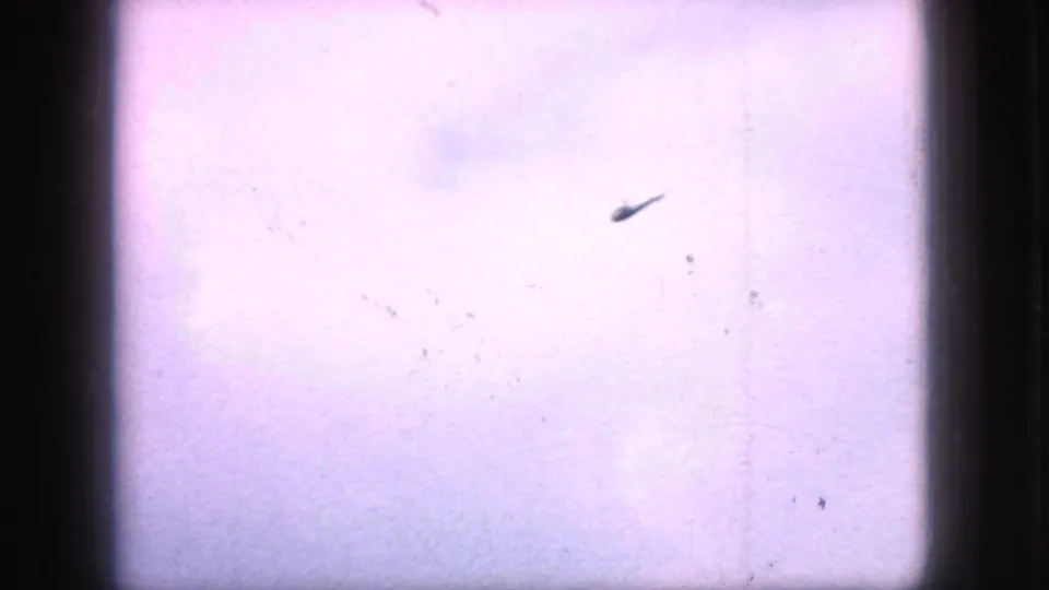 A distant helicopter-like silhouette crosses a pale sky in 1966 New Jersey 8mm footage, framed by dark gate edges and visible film dust.