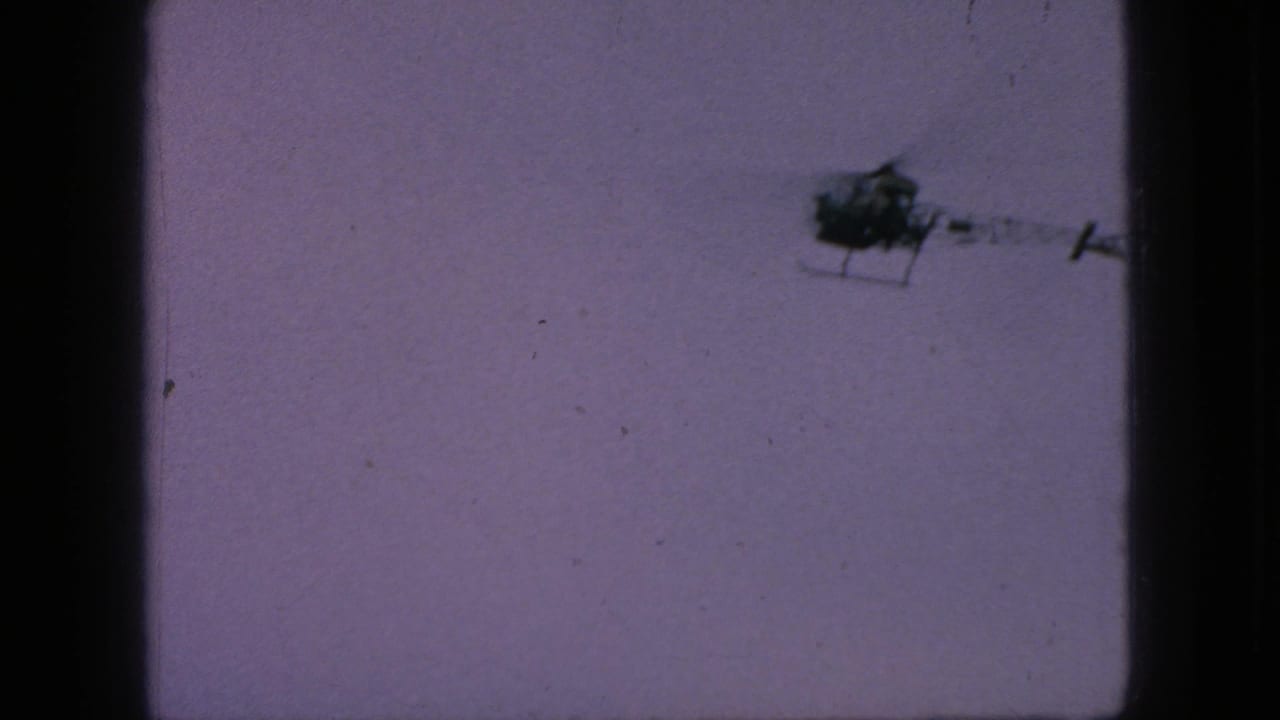 A small helicopter appears near the upper right of the frame, silhouetted against a muted sky in vintage 8mm footage from New Jersey.