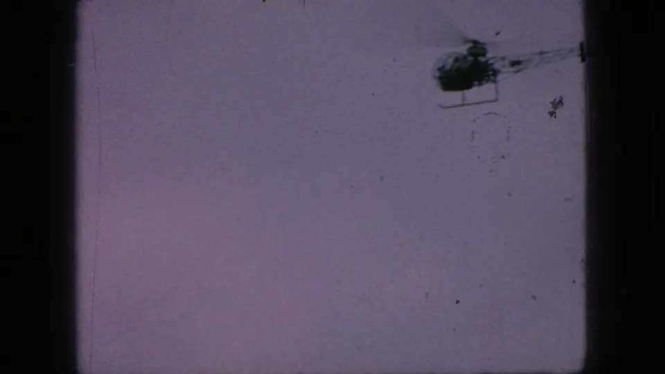 The helicopter is more centered in the upper frame as it moves across a flat, overcast sky, with dust and scratches visible in the 8mm scan.