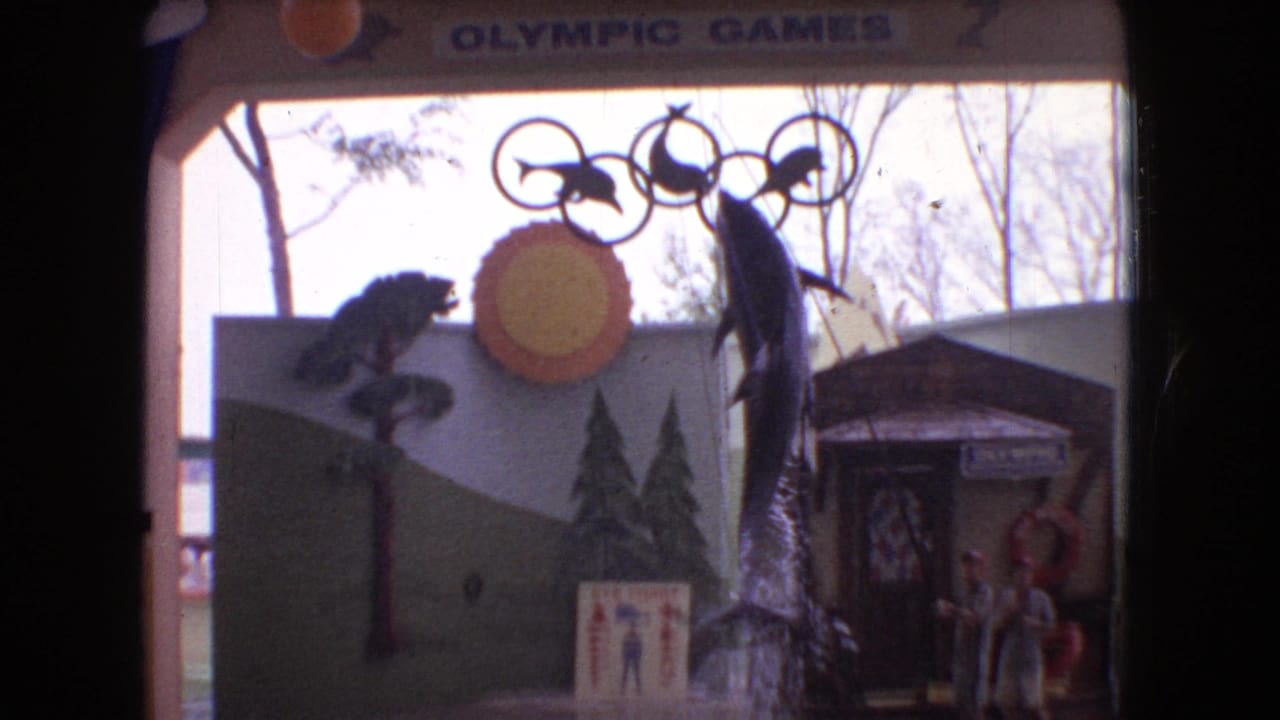 1966 Ohio 8mm frame showing a themed aquatic stage with a dolphin near the pool, painted backdrop panels, distant performers, and Olympic Games lettering above.