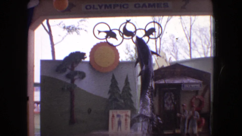 1966 Ohio 8mm frame showing a themed aquatic stage with a dolphin near the pool, painted backdrop panels, distant performers, and Olympic Games lettering above.