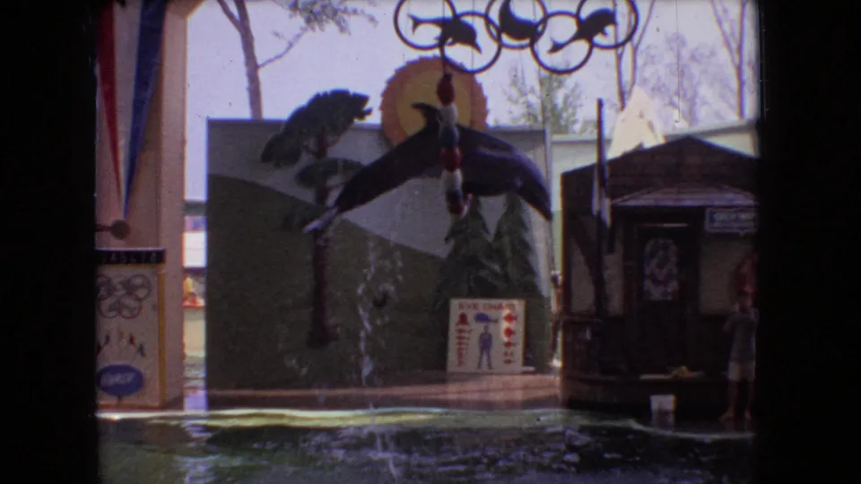 1966 Ohio 8mm frame of a dolphin leaping near hanging rings and show decorations, with a trainer visible on the platform beside the pool.