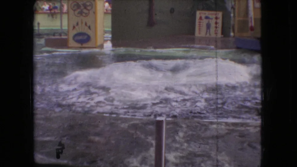Foam and rippling water spread across the aquarium performance pool after the jump, with signage, railings, and distant spectators in the background.
