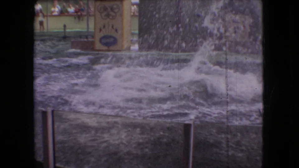 A broad splash spreads across the show pool after the dolphin movement, with railings, painted scenery, and background audience areas visible through the soft 8mm image.