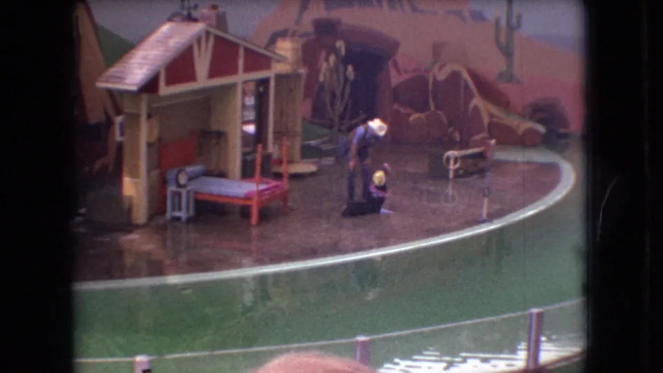 1966 Ohio home-movie frame showing the performer bending beside the seal during the act, with Western-themed props, painted cacti, railings, and audience heads at the frame edge.