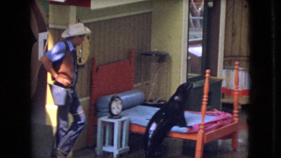 The 1966 Ohio 8mm frame shows the man facing the sea lion in a staged indoor setup with a red-framed bed, blue mattress, small side table, clock, and reflective floor.