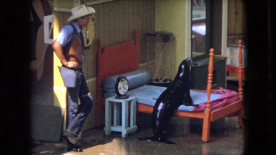 In the closing frame, the black sea lion rises beside the red-framed bed as the man in cowboy attire stands nearby in the 1966 Ohio home-movie scene.