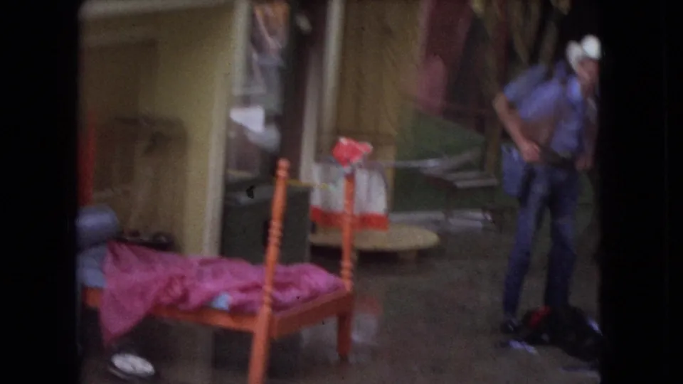 1966 Ohio home-movie frame showing a male animal trainer in a cowboy hat beside a small bed with pink bedding and painted exhibit scenery.