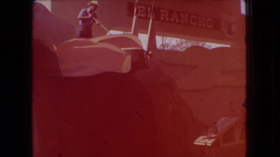 1966 Ohio 8mm frame showing a man in a hat working above a large colorful rooftop figure near a partially visible El Rancho sign, with strong red color shift and film borders.