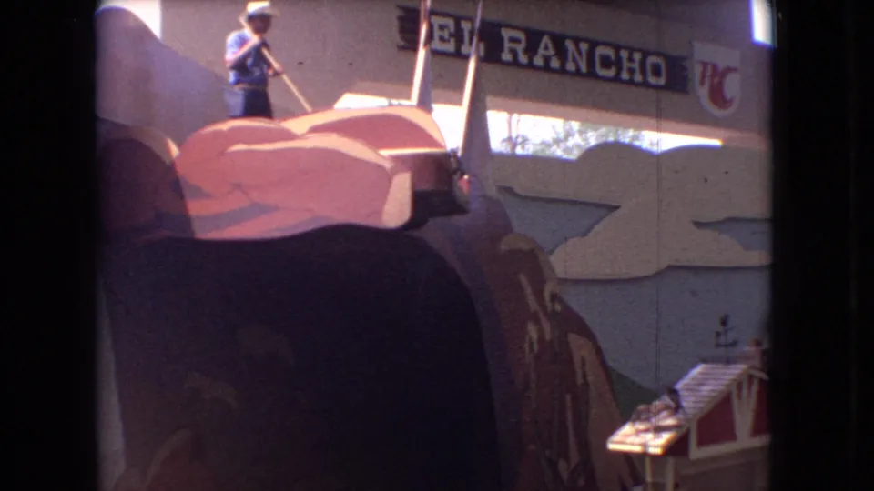 1966 Ohio 8mm frame showing a man in a hat washing or working on a large colorful rooftop figure beside the El Rancho sign and a restaurant facade.