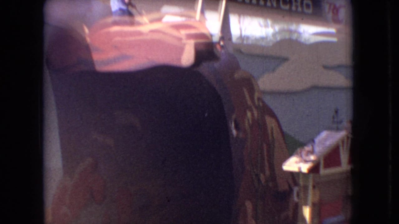 1966 Ohio 8mm frame showing a visitor's view of a man-made theater attraction with painted cliff scenery, partial signage, and an audience member's head at the bottom edge.