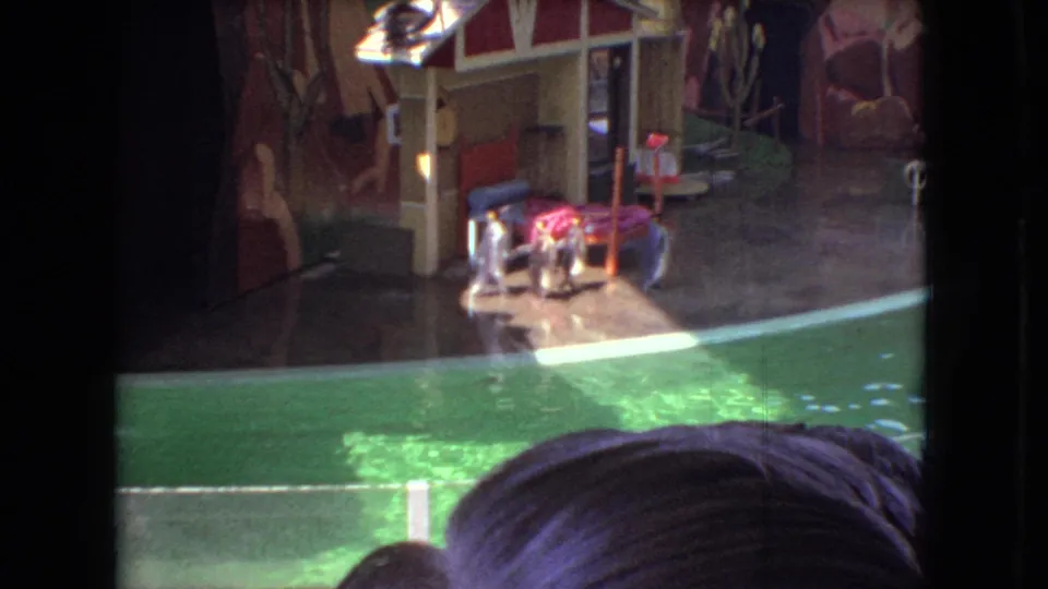 1966 Ohio 8mm frame showing penguins near a wet platform beside a green pool, with a small stage hut, painted scenery, and audience heads framing the lower edge.