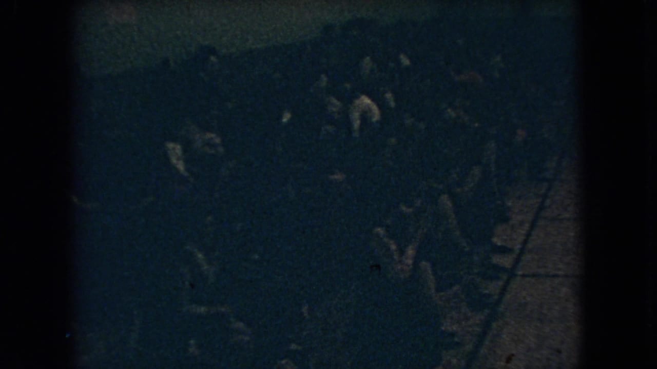 Dim crowd scene with several standing figures and indistinct movement in a room.