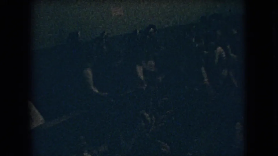 1966 Ohio 8mm frame showing a low-exposure overhead view of a crowded indoor celebration, with figures reduced mostly to silhouettes and light clothing highlights.