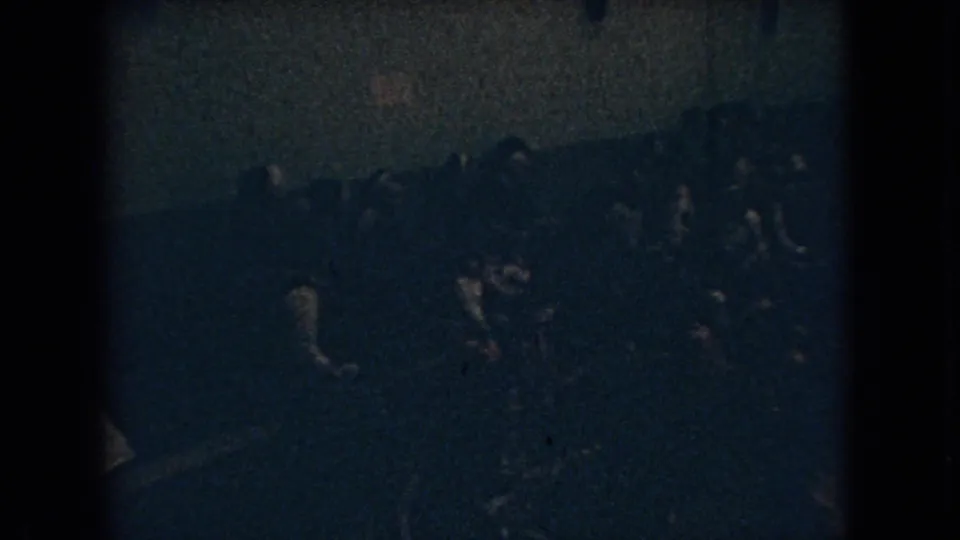 1966 Ohio 8mm frame showing a dense group of people in a dim private interior, seen from above with heavy grain and indistinct faces.