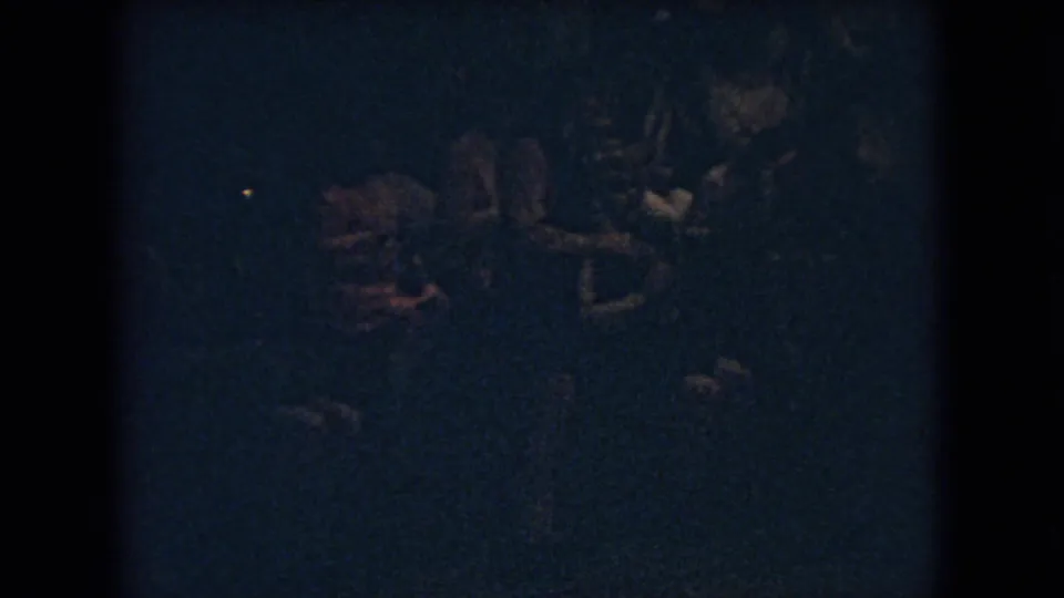 The 1966 Ohio home-movie frame remains heavily underexposed, with a few spectators or players faintly visible along the edge of nighttime basketball action.