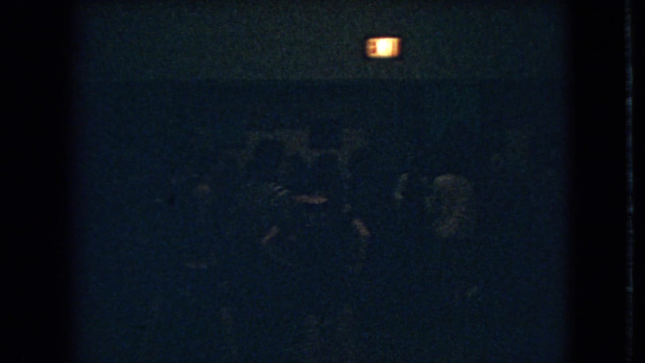 Dim 1966 Ohio 8mm frame showing shadowed people gathered below a glowing exit sign in a dark interior.