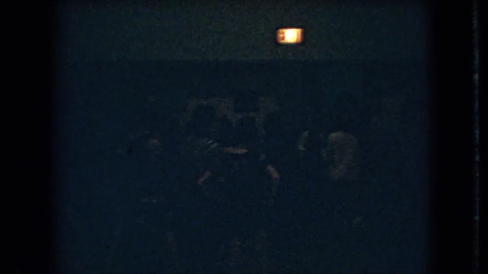 Dim 1966 Ohio 8mm frame showing shadowed people gathered below a glowing exit sign in a dark interior.