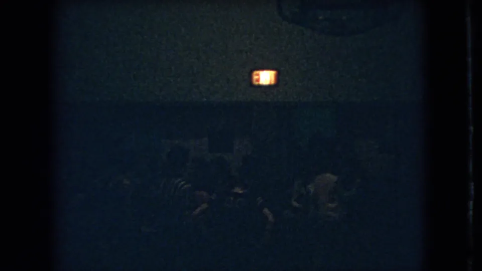 Low-light Ohio 1966 home-movie frame with silhouetted adults moving or standing near an illuminated exit sign.