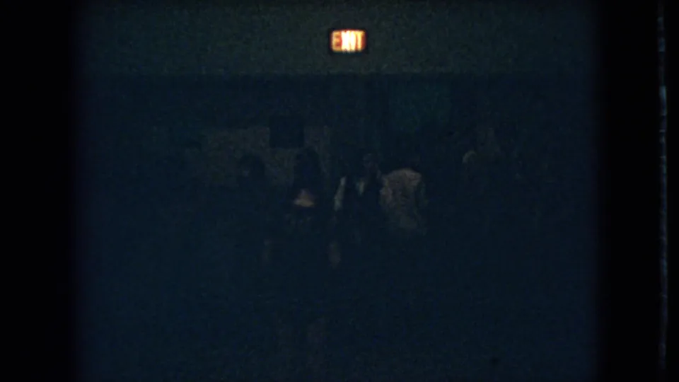 Grainy 1966 Ohio frame showing several dark figures in a poorly lit interior beneath a glowing exit sign.